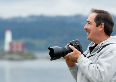 Man smiling holding a camera while outside by Fisgard Lighthouse in Victoria BC